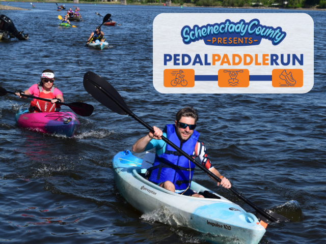 imagine of a male kayaker in a blue kayak and life vest paddling the Mohawk River in Niskayuna followed by a female kayaker in a pink kayak and life vest.  The Pedal Paddle Run Logo is situated in the top right corner.