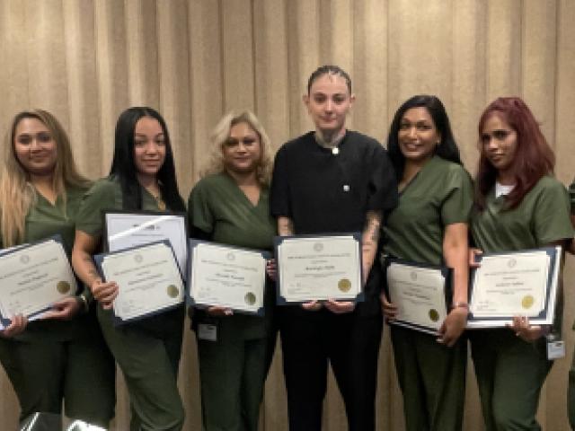 Photo of all eight CNA graduates holding certificates, County Legislature Chair Hughes, and their trainer