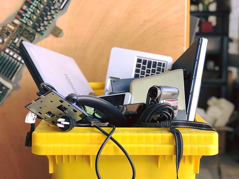 A yellow bin filled with discarded electronic devices including cellphones, laptops, camcorders, external hard drives, power cables, and plugs. 